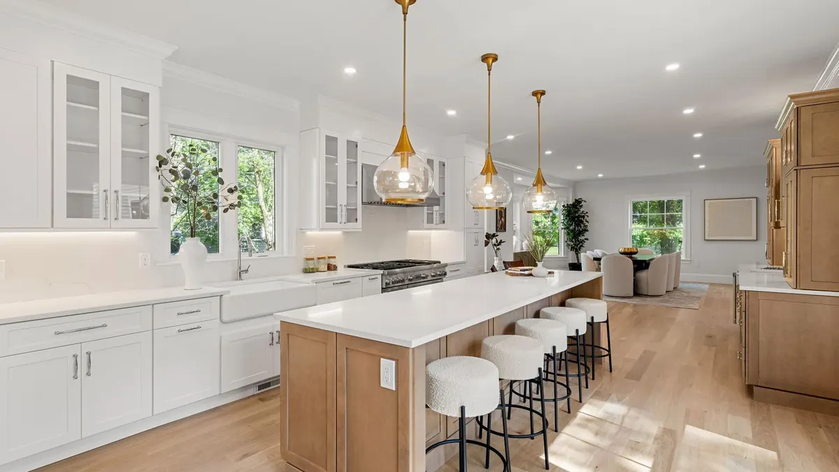 A warm white-and-wood kitchen with a natural-oak island, brass pendants, and glass-front cabinetry open to the living room — a Bay State Kitchen Gallery project in Newton