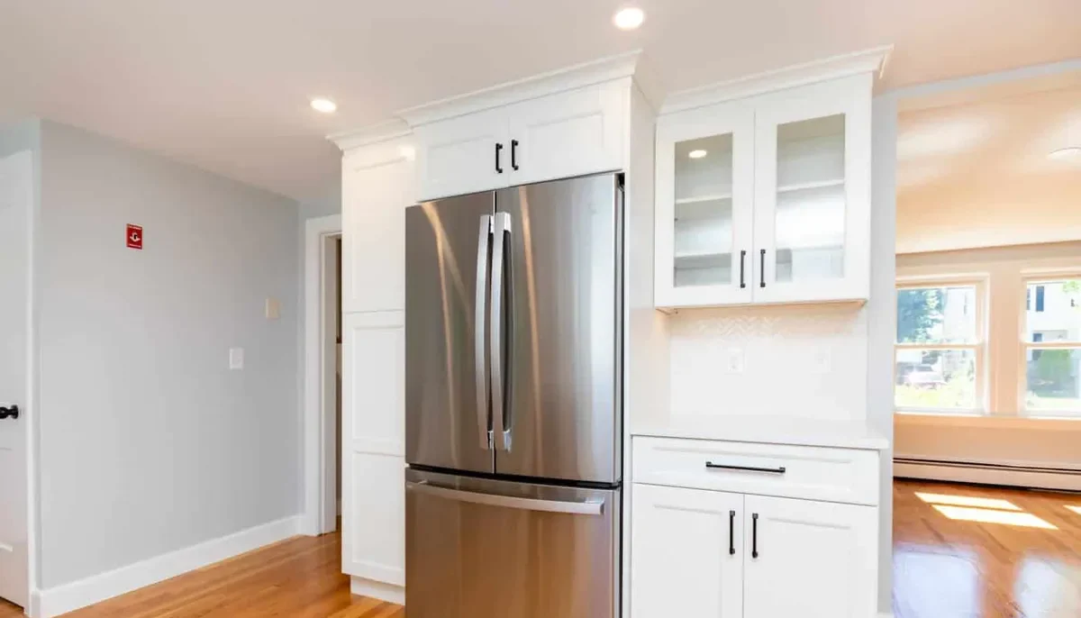 Built-in stainless refrigerator flanked by white shaker cabinets and a glass-front hutch in this Waltham kitchen