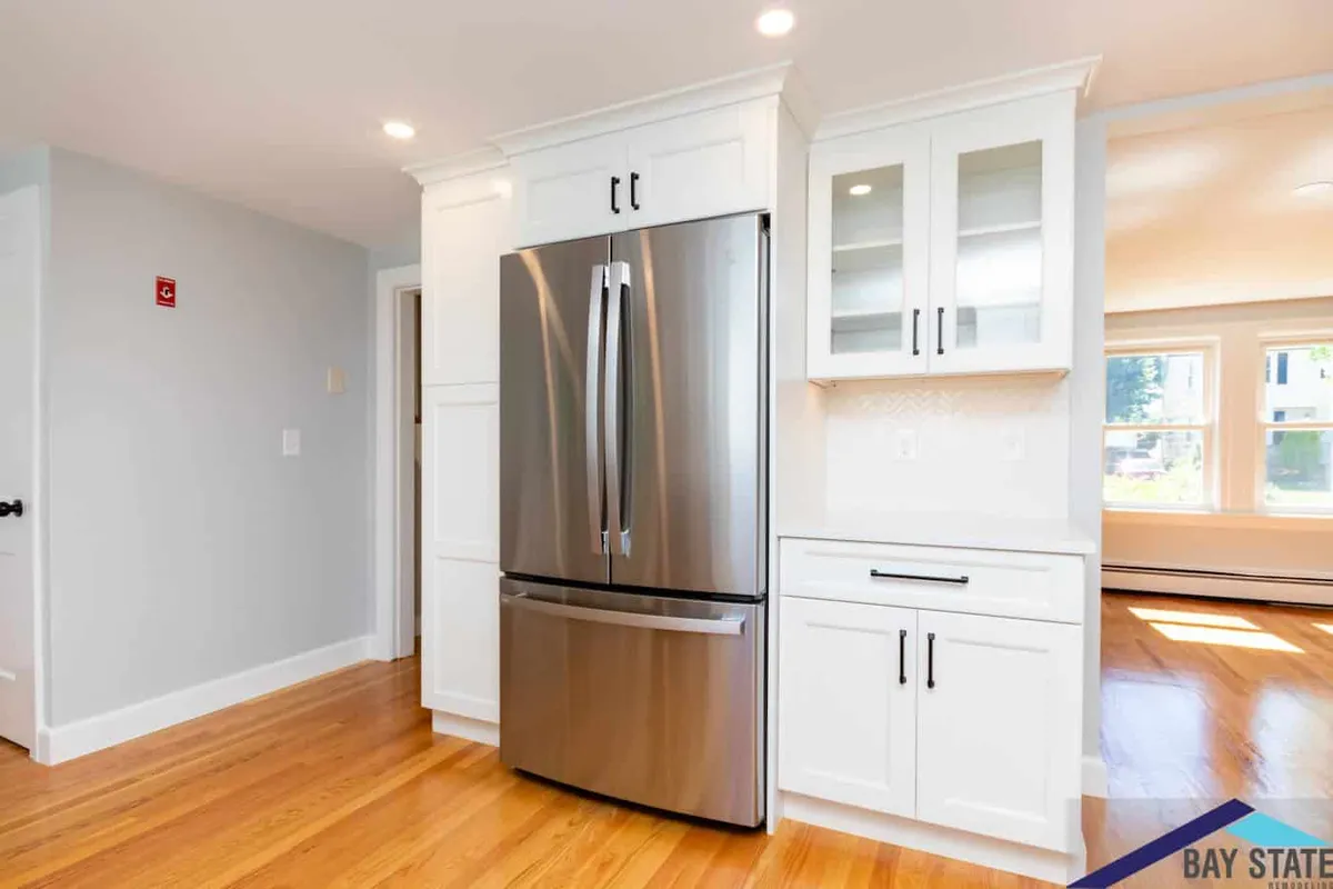 Built-in stainless refrigerator flanked by white shaker cabinets and a glass-front hutch in this Waltham kitchen