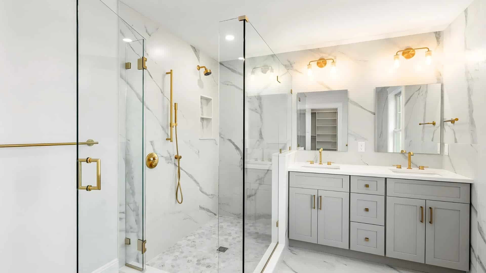A finished bathroom with marble-look tile, a frameless glass shower, brushed-gold fixtures and a gray double vanity on Auburndale Ave. in Newton