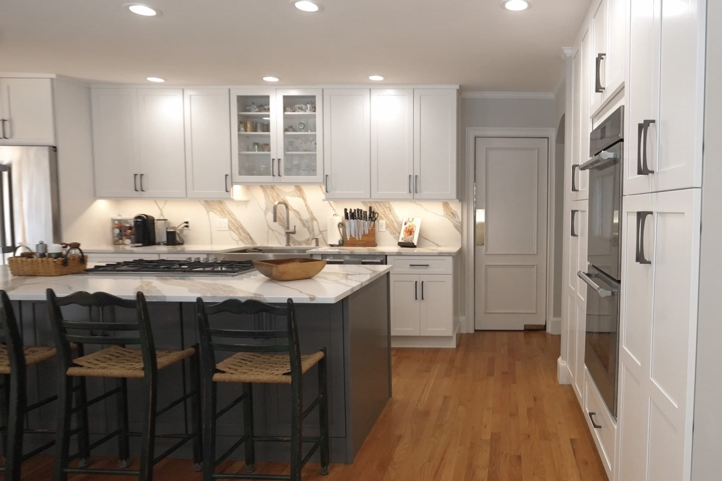 A finished white shaker kitchen with a marble backsplash, glass-front uppers and a charcoal island with rush-seat stools on Brook Trail Rd. in Wayland