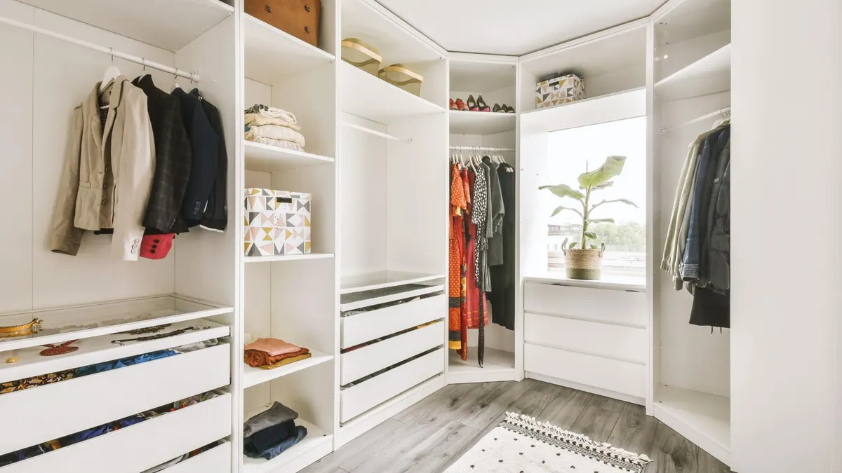 Corner walk-in closet with white built-in shelving, hanging rods, drawers, and a window with a plant